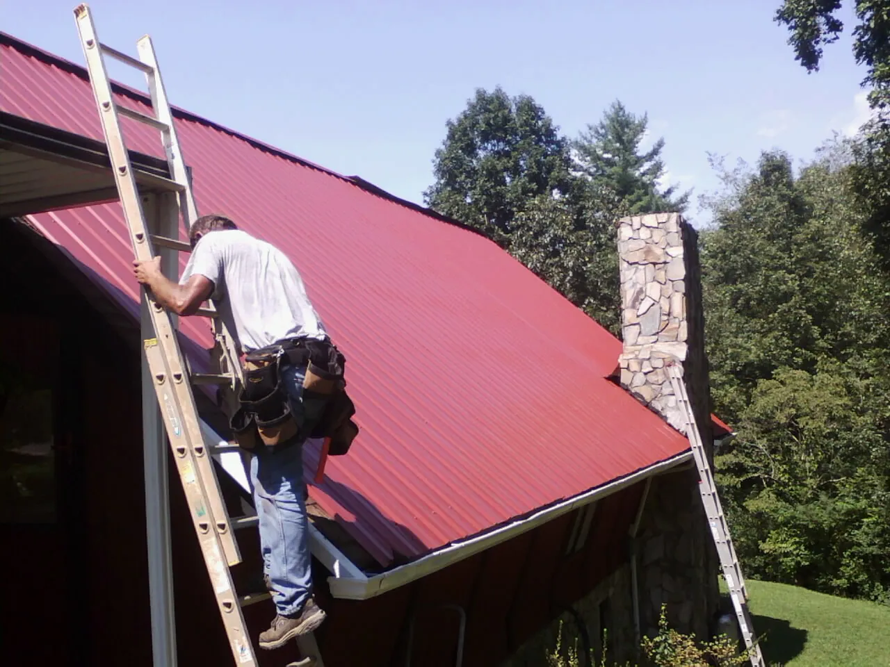 Roofer on a ladder beside a red metal roof and stone chimney in Clarksburg